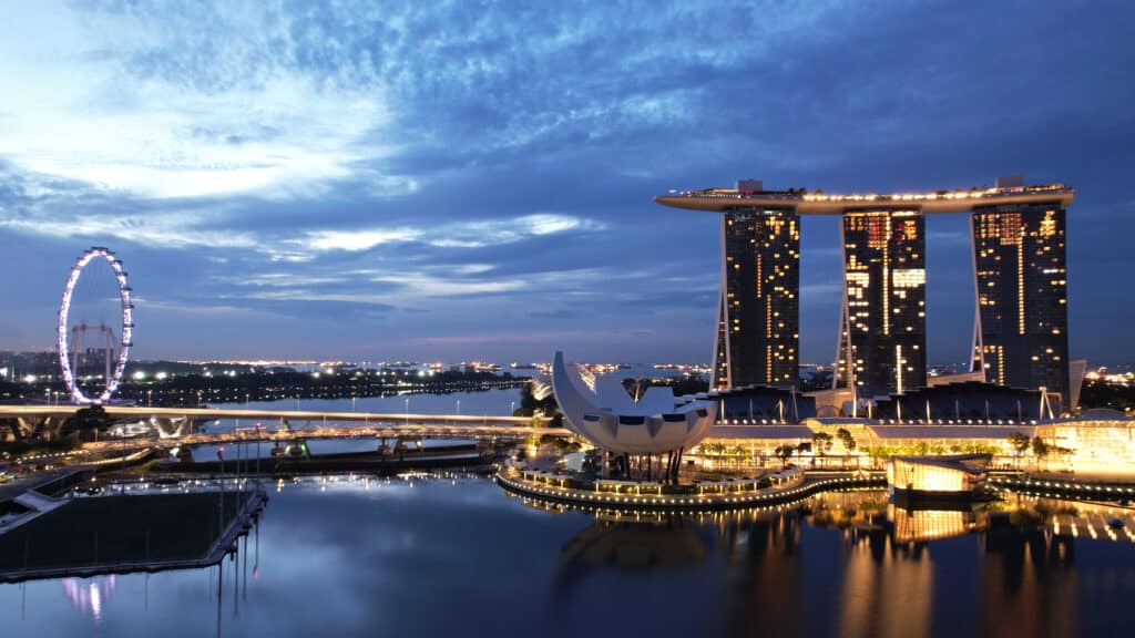 City skyline at dusk with illuminated ferris wheel and futuristic buildings reflecting on water.