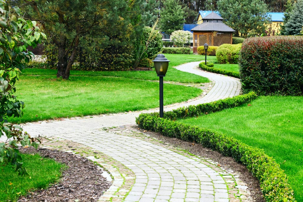 Winding garden path with lush greenery and bushes, leading to a wooden gazebo under a blue sky.