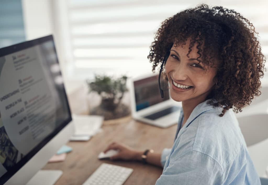 Smiling woman with headset working on computer, providing customer support from her home office.