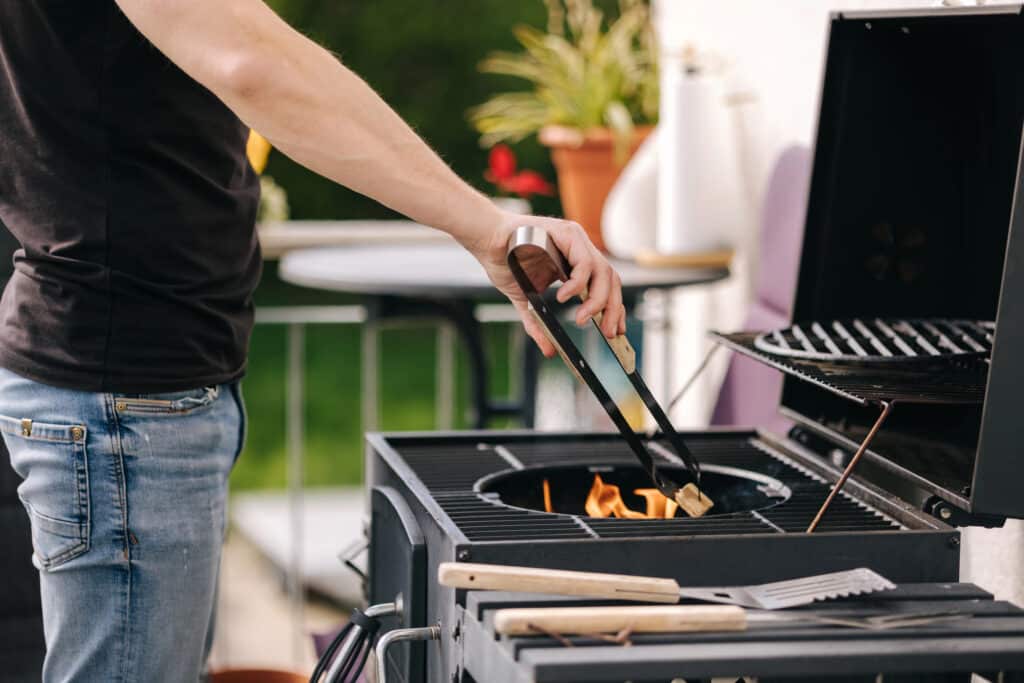 Person grilling on a barbecue with tongs and tools, outdoor kitchen setting, summer cookout.