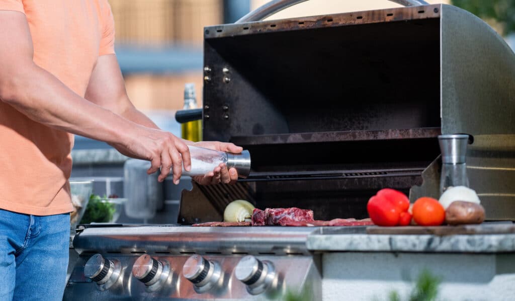 Person seasoning meat on an outdoor grill with vegetables nearby.