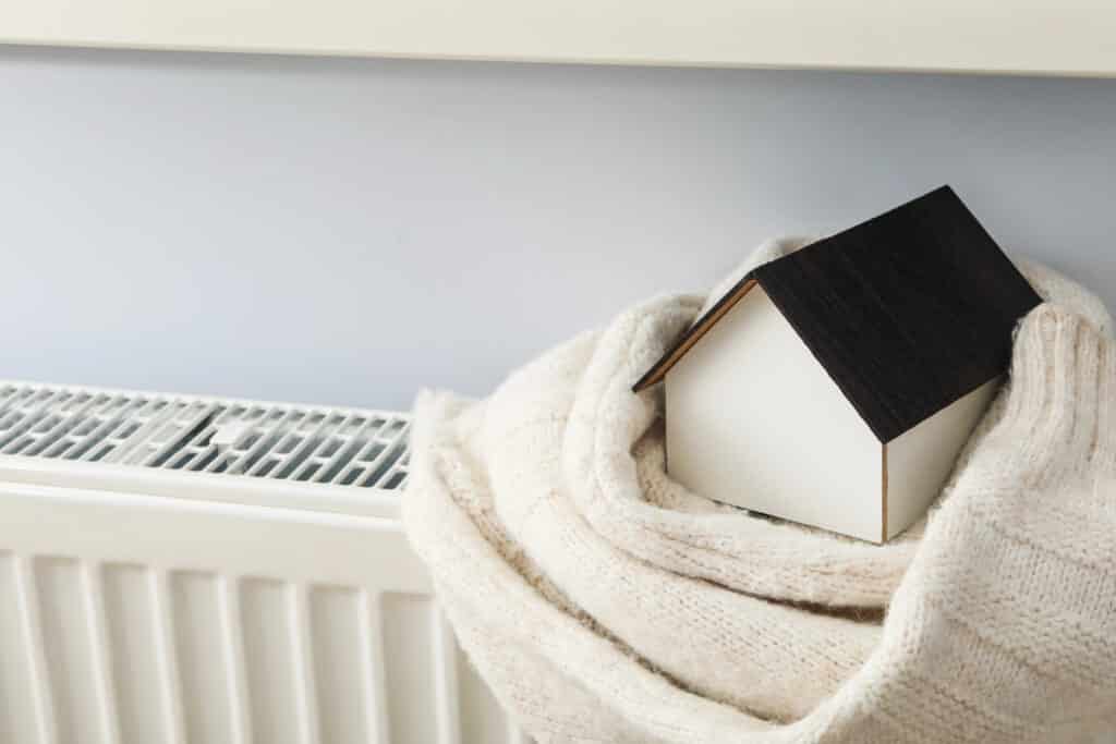 Depositphotos_678126976_XL Mini house wrapped in a warm blanket beside a radiator, symbolizing home heating and comfort during winter. | Sky Rye Design Mini house wrapped in a warm blanket beside a radiator, symbolizing home heating and comfort during winter.
