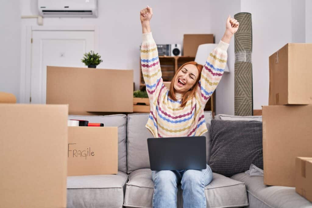 Woman happily celebrating moving day on a couch surrounded by boxes, using a laptop.