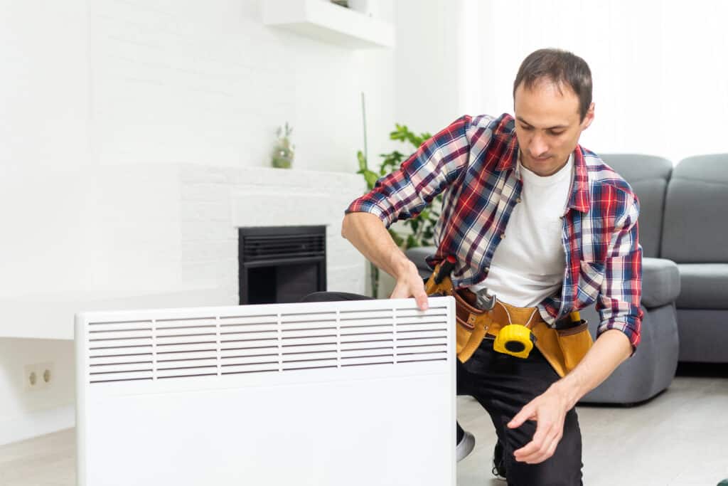 Man in plaid shirt installing white heater in living room, wearing tool belt.