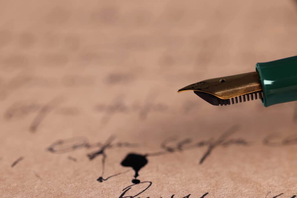 Close-up of fountain pen writing on antique paper, with ink splatters and cursive script in the background.