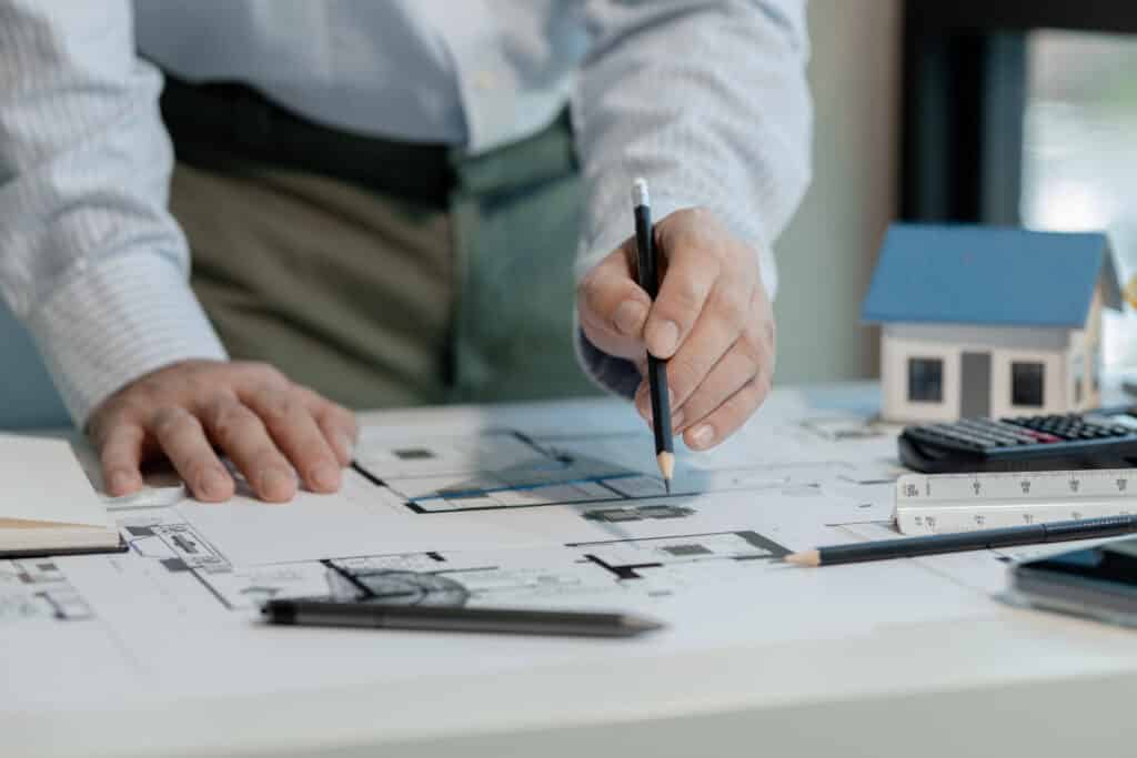 Architect working on blueprints with a pencil, ruler, and miniature house model on a desk.