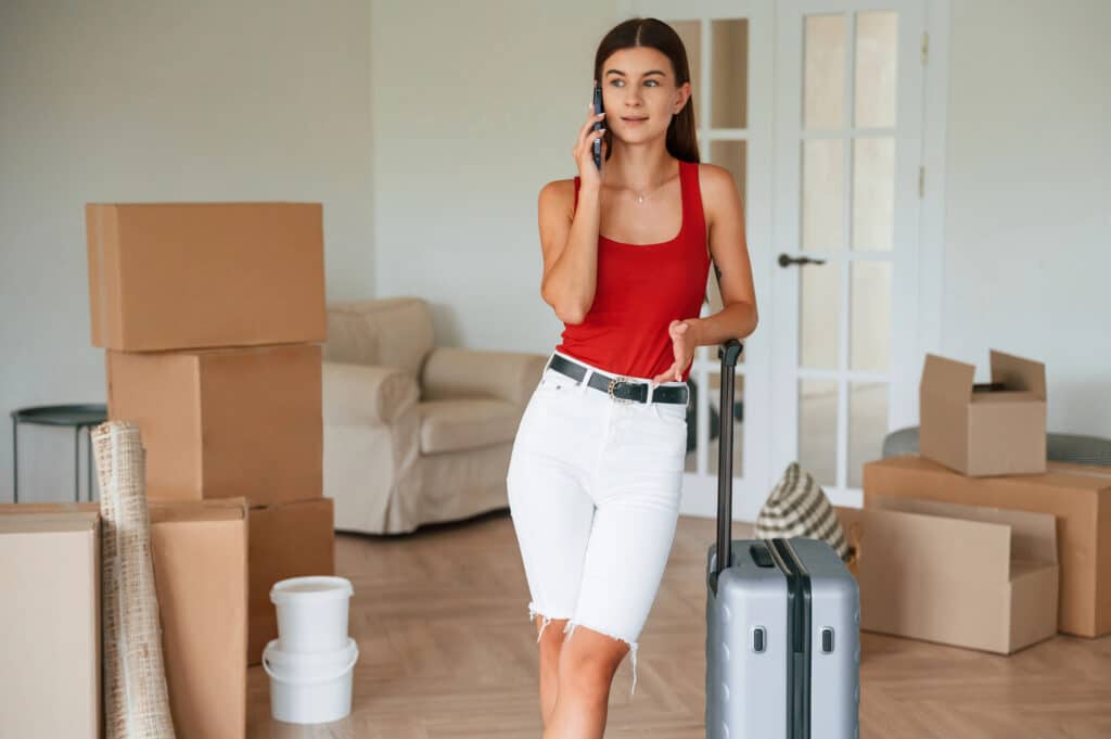 Depositphotos_791457542_XL Woman talking on phone, standing with suitcase and boxes in room. | Sky Rye Design Woman talking on phone, standing with suitcase and boxes in room.
