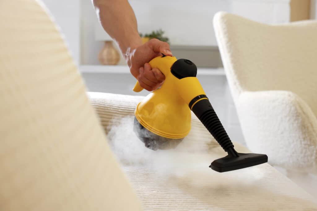 Hand using a yellow steam cleaner on a beige fabric couch for efficient deep cleaning.