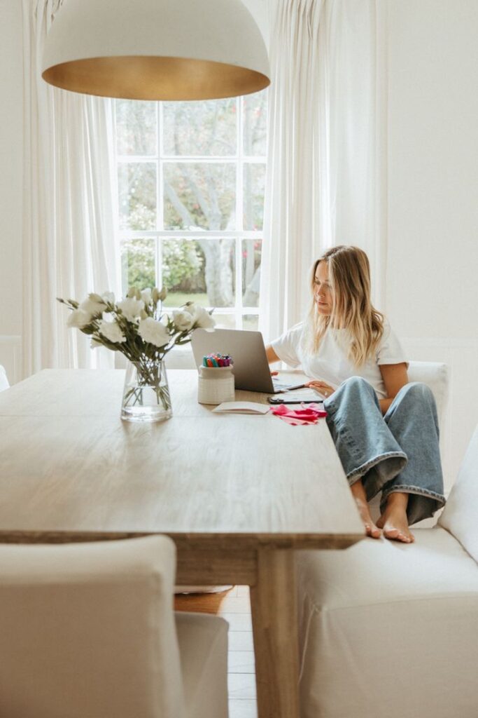 Woman working on a laptop at a cozy home office with flowers and a large window in the background.