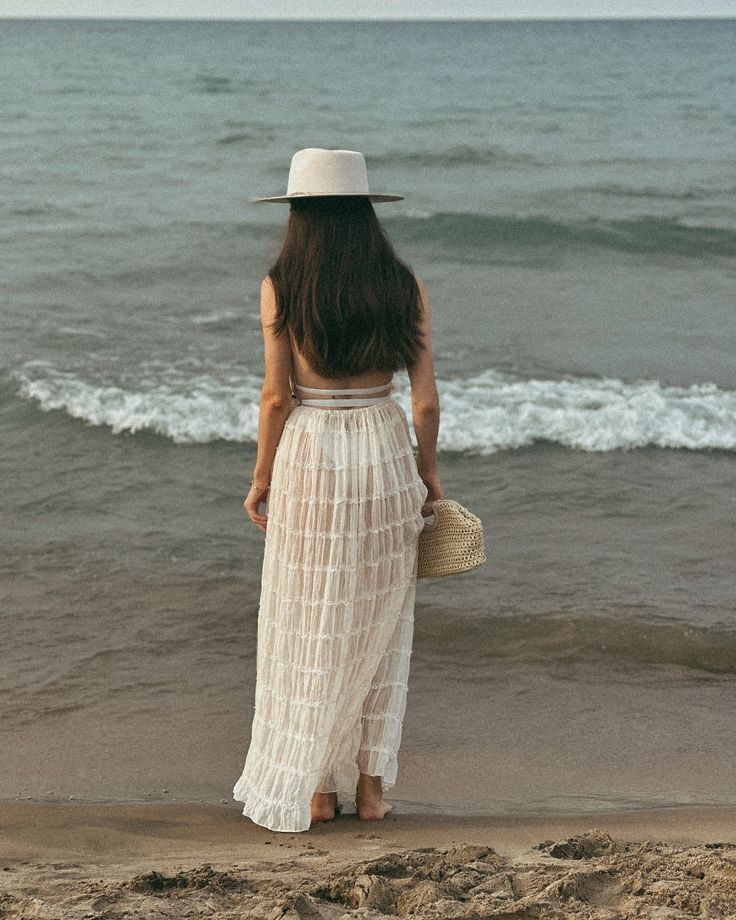 Woman in white dress and hat stands on beach, gazing at ocean waves.