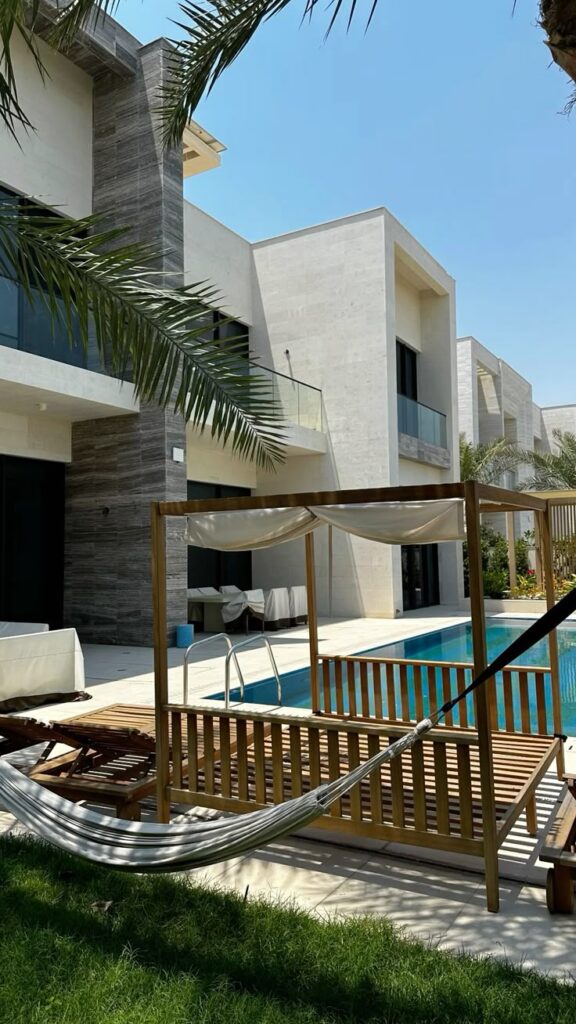 Modern luxury villa with pool, patio, hammock, and palm trees under clear blue sky.