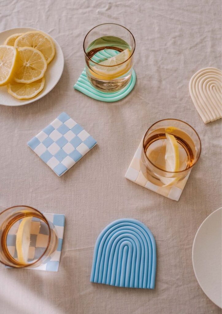 Glasses with lemon slices on colorful coasters and a plate of lemons on a neutral tablecloth.
