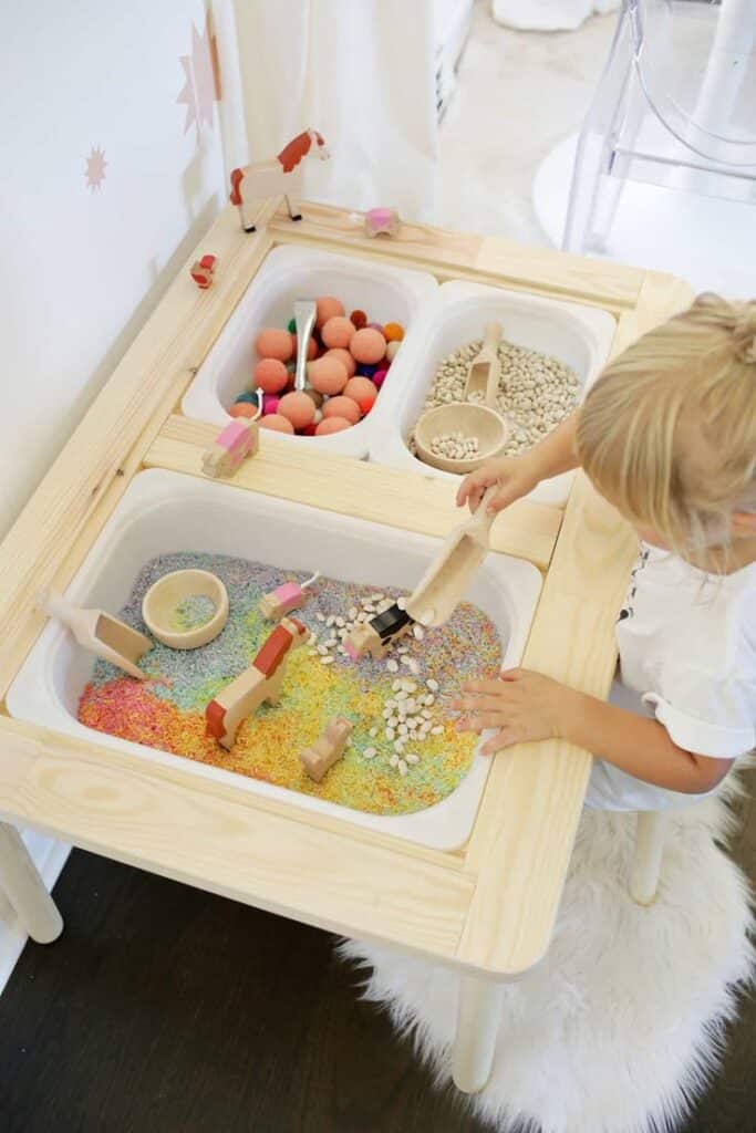 Child playing with colorful sensory table filled with beans, pom-poms, and toy animals for hands-on learning.