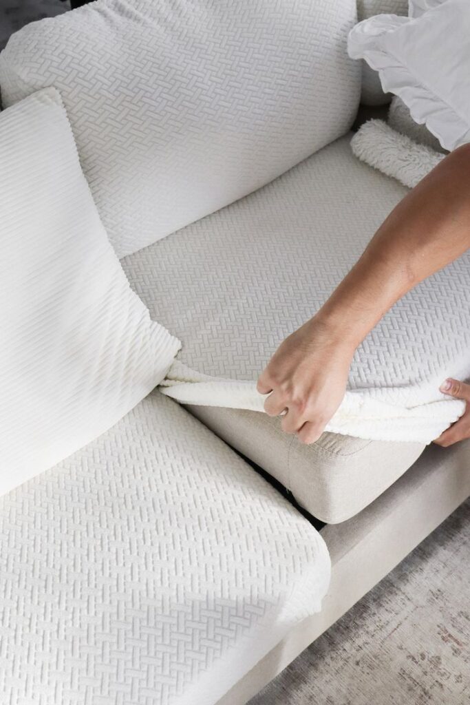 Person adjusting a white slipcover on a sofa cushion, enhancing the living room decor with a fresh, clean look.