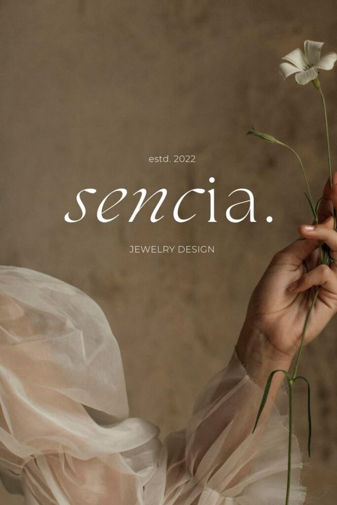Elegant hand holding flower with sencia text, jewelry design brand established in 2022, soft fabric background.