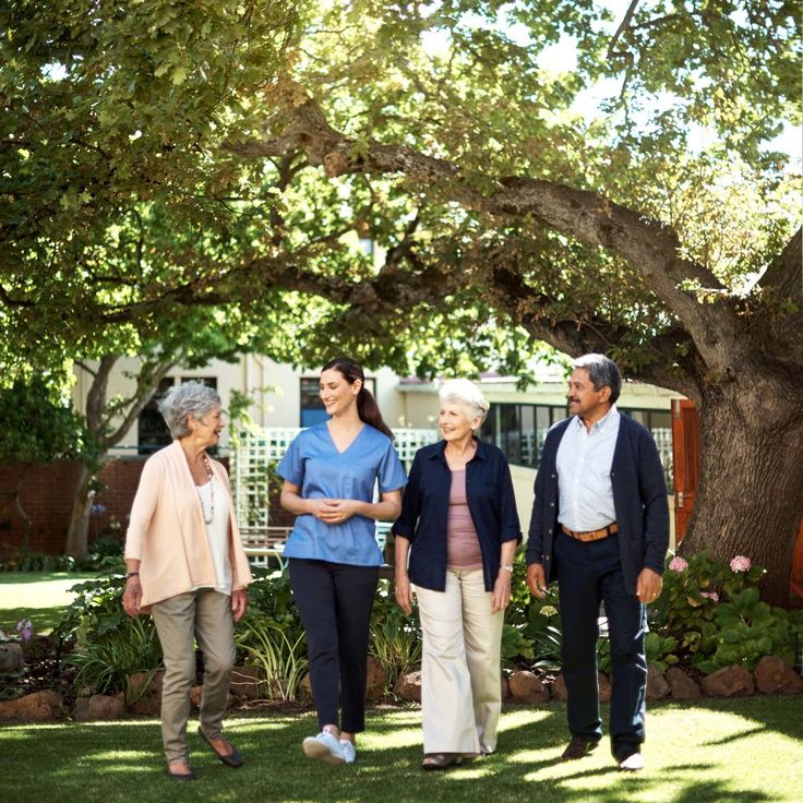 Group walking in a garden under a tree, including a caregiver in blue and three elderly people enjoying the outdoors.