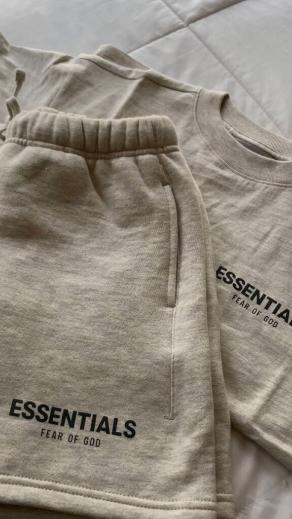 Beige Fear of God Essentials sweatshirt and shorts set on a bed, showcasing brand logo for stylish casual wear.