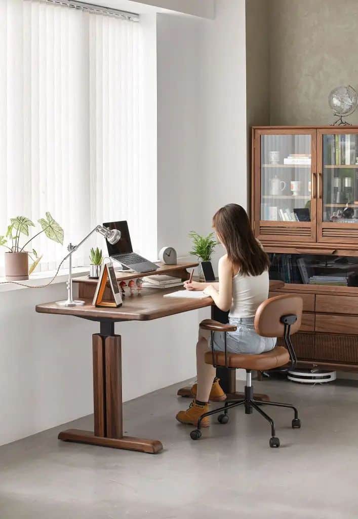 Woman working at a home office desk with plants, laptop, and shelves in a modern, well-lit room.
