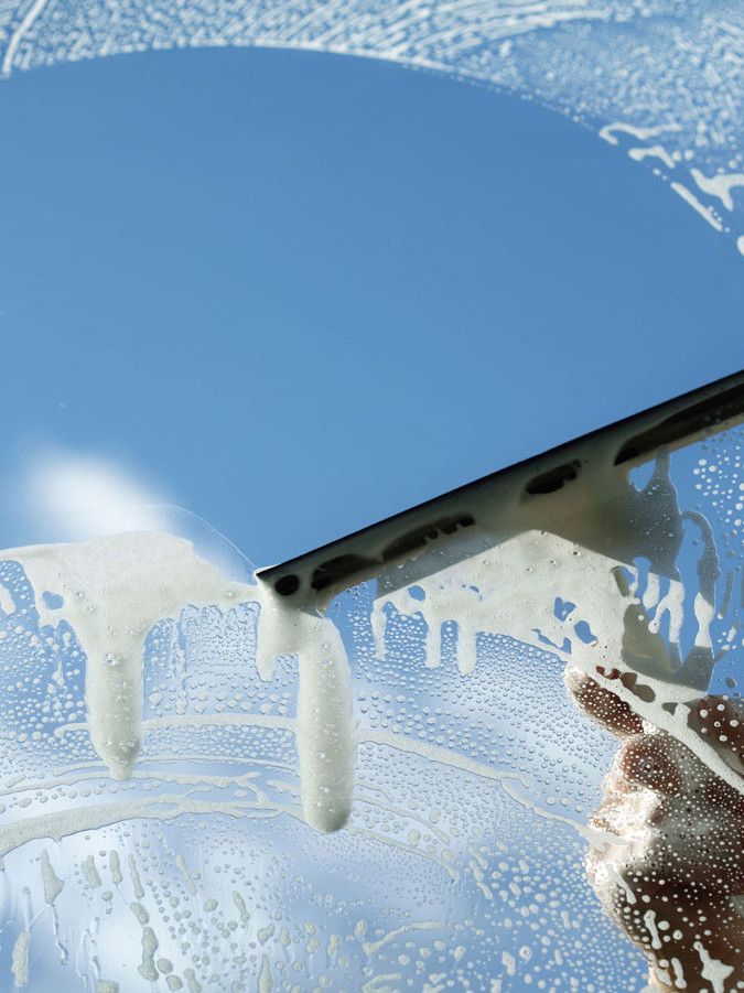 Hand using a squeegee to clean soapy window, revealing clear blue sky background.