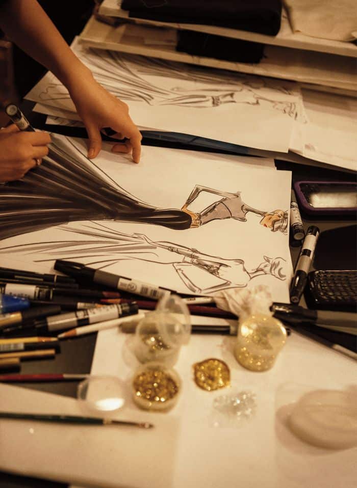 Fashion designer sketching a gown surrounded by pens and glitter on a creative workspace.