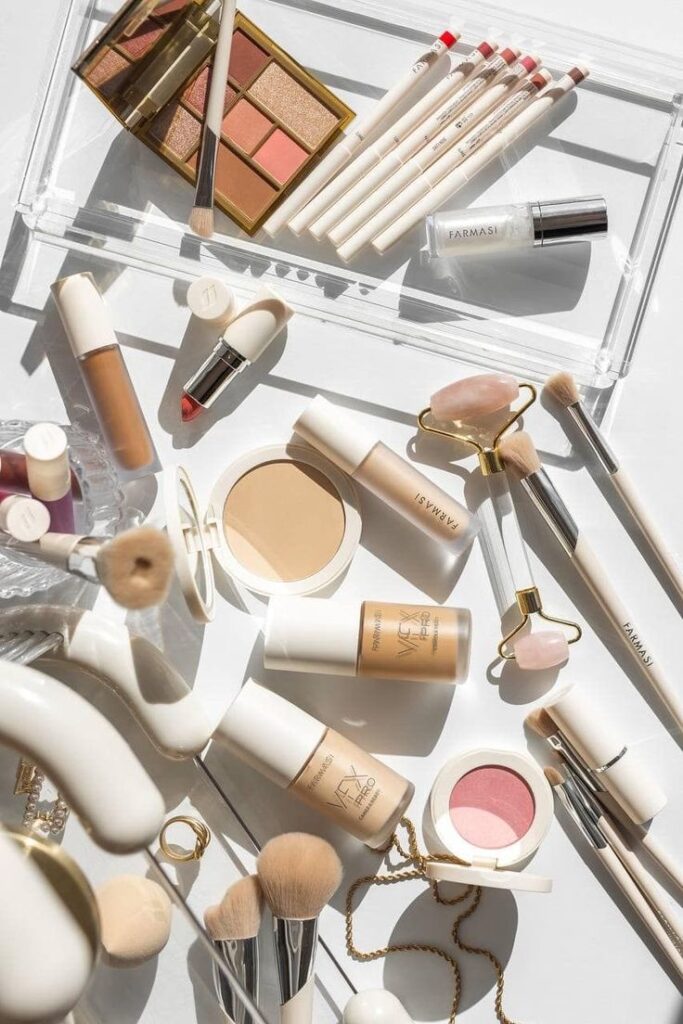 Assorted makeup products and brushes on white background, featuring palettes, lipsticks, and foundation bottles.