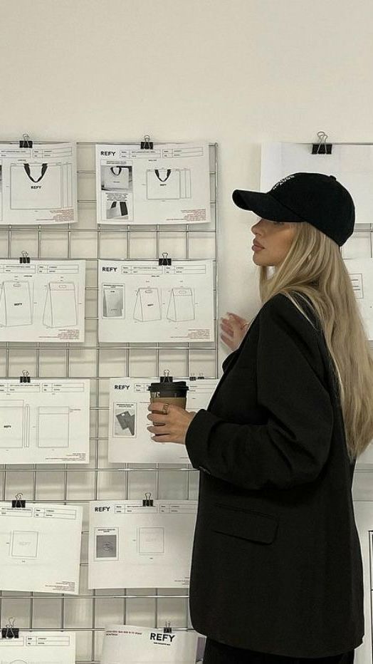 Woman in black cap examines design sketches on wall, holds coffee cup.