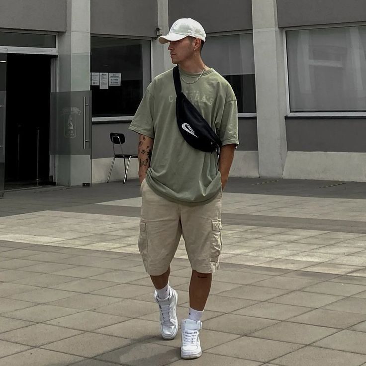 Casual streetwear outfit with cap, oversized t-shirt, shorts, and sneakers on a city sidewalk.
