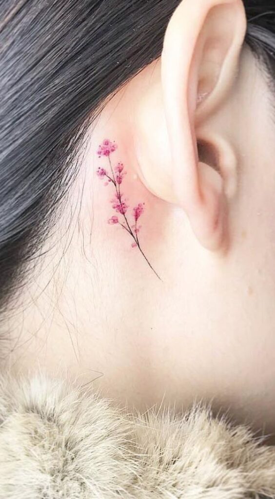 Delicate pink floral tattoo behind the ear on a person with dark hair.