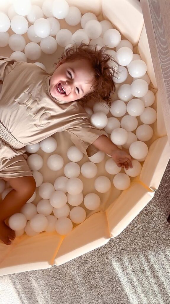 Smiling child playing in a ball pit filled with white balls, wearing a beige outfit, enjoying a fun-filled moment.