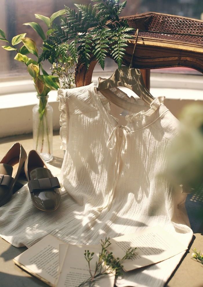 White blouse on hanger with loafers, books, and greenery in soft sunlight, creating an elegant, cozy atmosphere.
