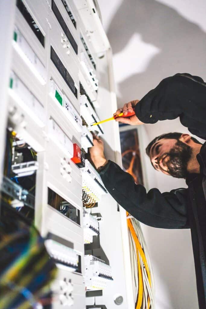 Electrician working on a control panel with yellow screwdriver, adjusting electrical components, smiling.