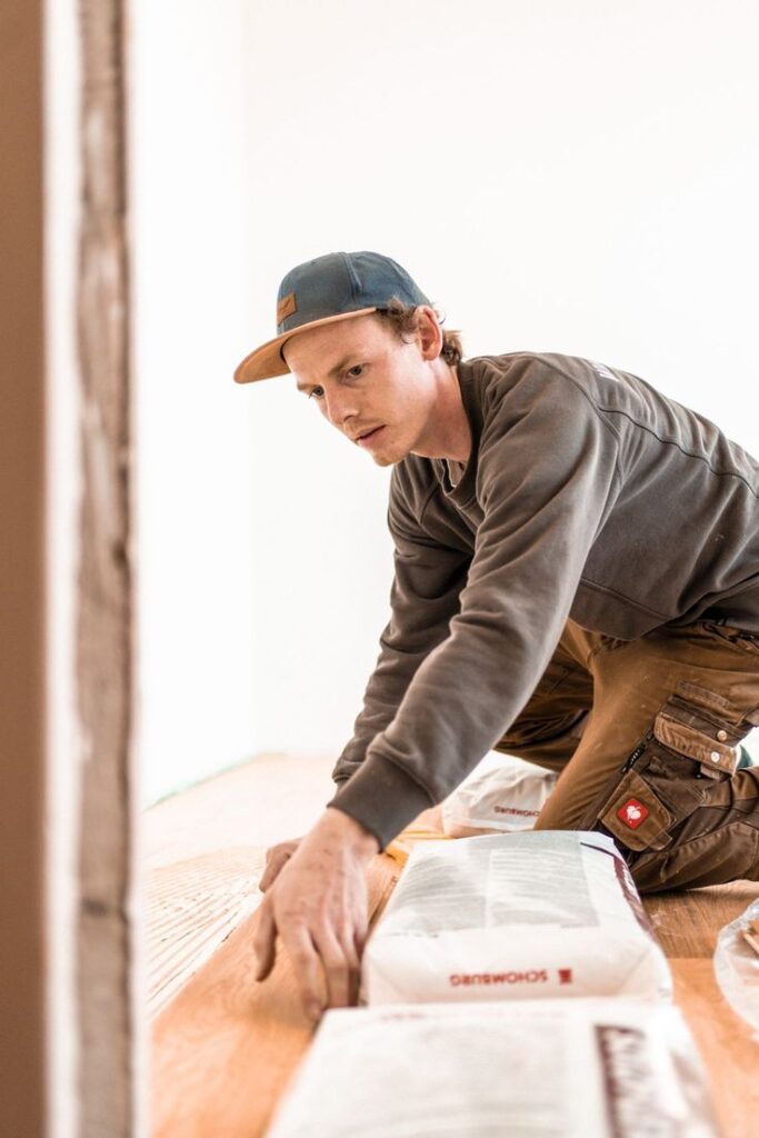 Worker installing wooden flooring in a room, wearing a cap and brown pants. Construction and renovation concept.
