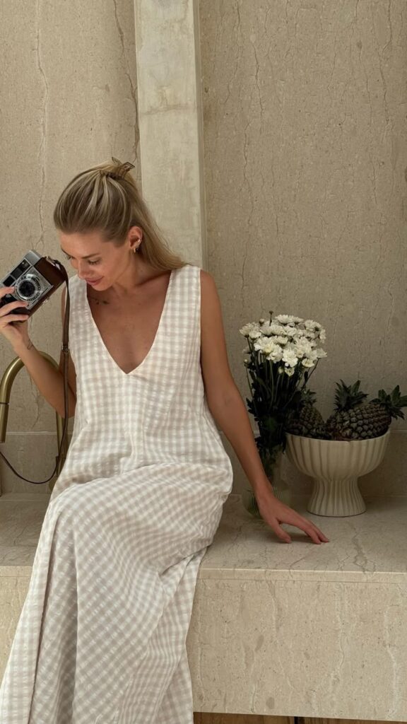 Person in checkered dress with vintage camera, sitting near flowers and pineapples on a stone porch.