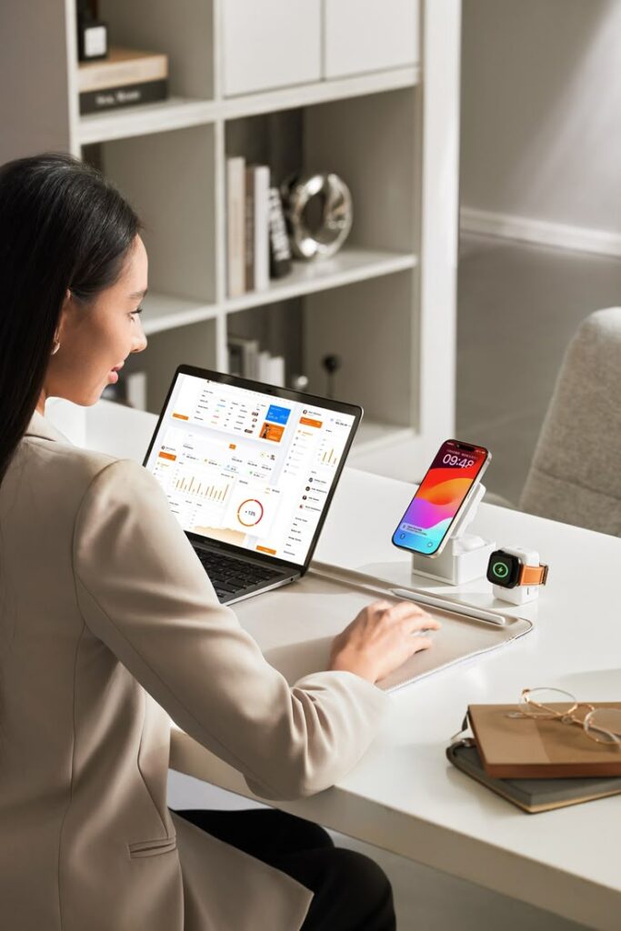 Woman working on a laptop with charts, phone, and smartwatch on desk in modern office setting.