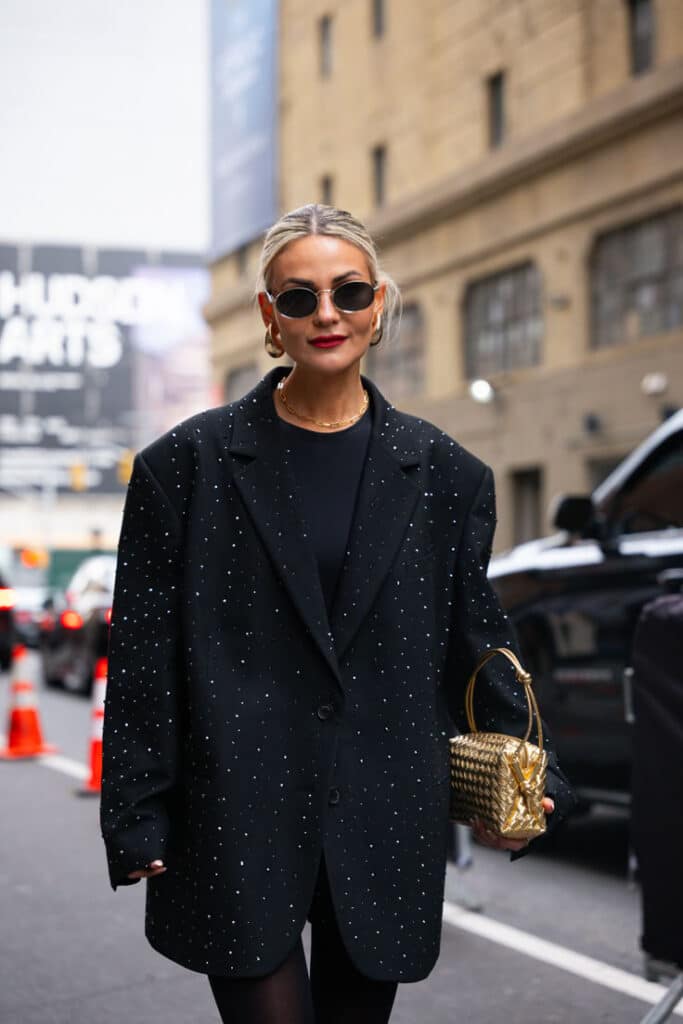 Fashionable woman in sparkly black blazer and sunglasses, holding a gold handbag on a city street.