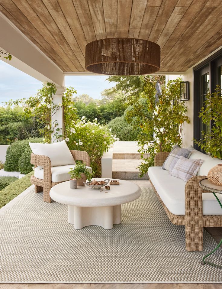 Cozy outdoor patio with wicker furniture, cushions, a round table, and lush greenery under a wooden ceiling.