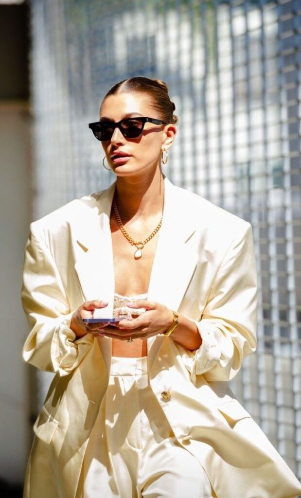 Stylish woman in a cream suit and sunglasses, holding a phone with a confident look near a metallic fence.