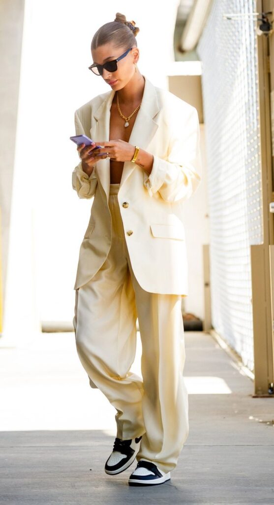 Fashionable woman in cream suit and sunglasses, walking and checking phone outdoors in natural light.