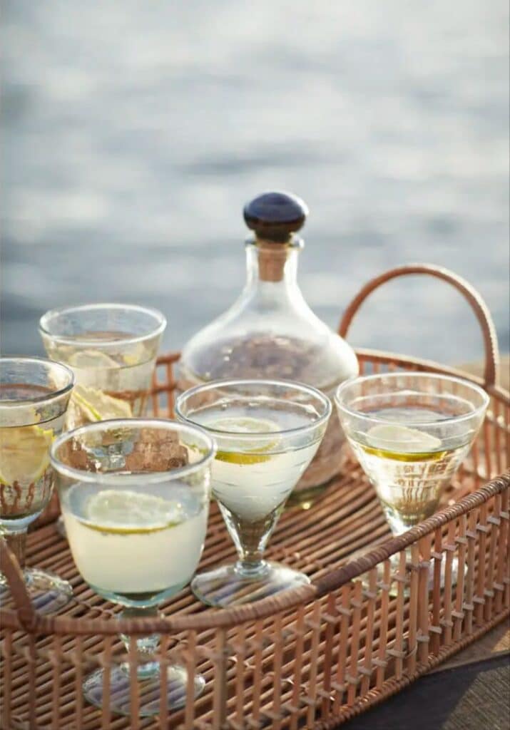 Wicker tray with cocktails and a decanter by the seaside; refreshing drinks garnished with lemon slices.