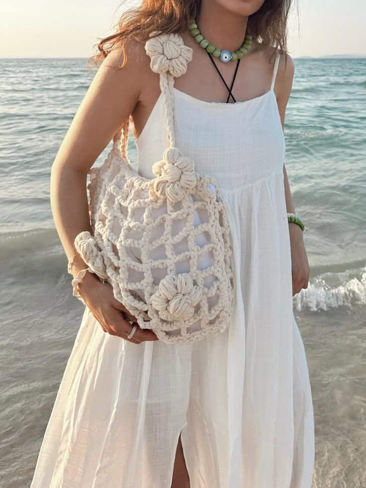 Woman in white dress with crochet bag by the ocean at sunset.