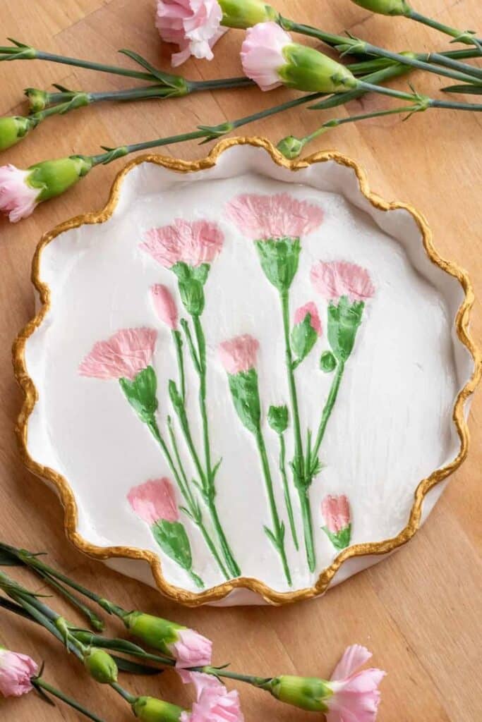 Decorative plate with pink floral design and gold edges surrounded by fresh pink carnations on a wooden surface.