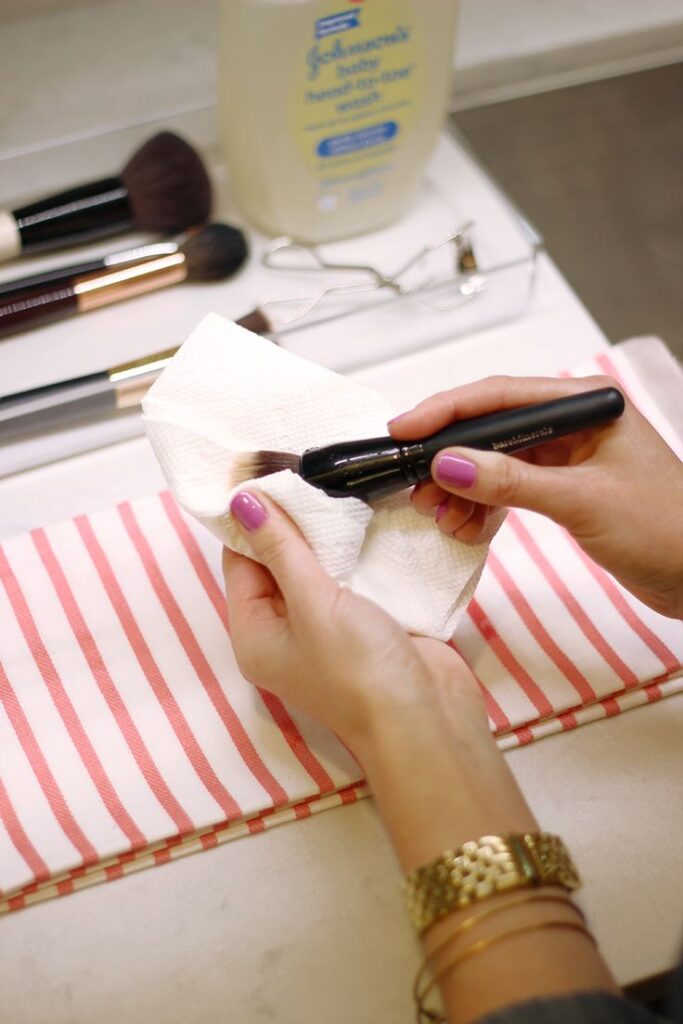 Cleaning makeup brushes with a paper towel, surrounded by tools and cleanser on a striped cloth.