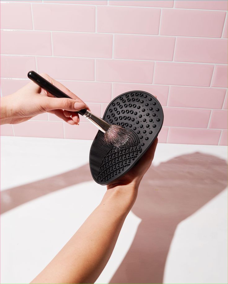 Makeup brush being cleaned on a textured silicone pad against a pink tile background.