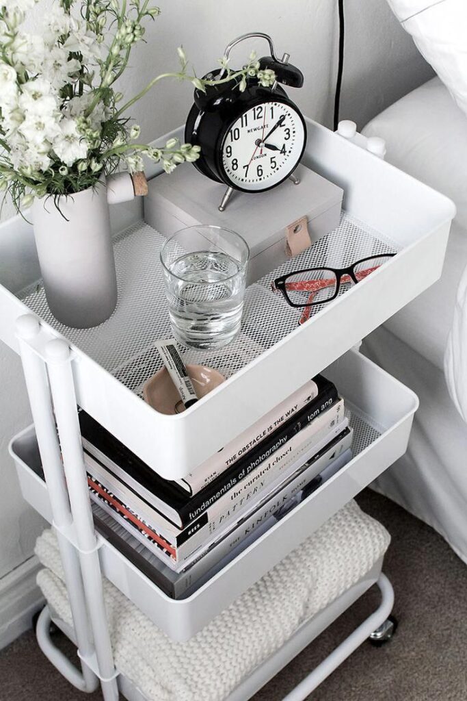 Bedside cart with alarm clock, glass of water, books, glasses, and floral decor for a stylish bedroom setup.