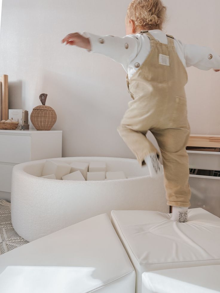 Child in beige overalls jumps into white foam pit in a cozy room with wicker decor and soft lighting.