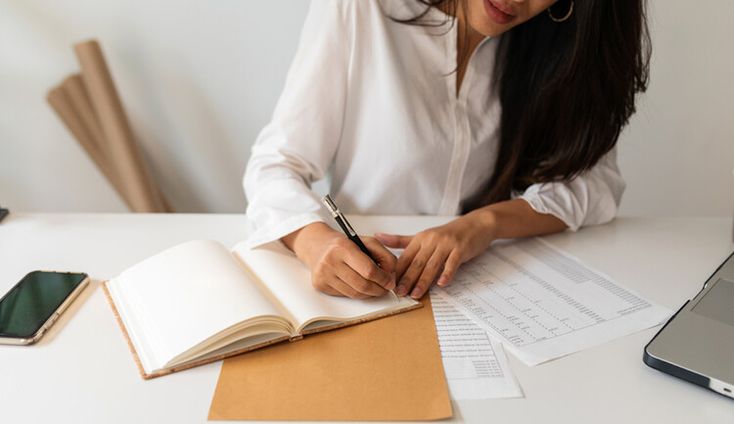I Thought I Had To Choose Between Paying Off Debt and Saving for RetirementBut Finance Pros Gave Me a Plan for Doing _Both_ Well+Good Woman writing in notebook at desk with documents and laptop nearby. | Sky Rye Design Woman writing in notebook at desk with documents and laptop nearby.