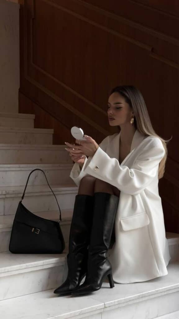 Stylish woman in white blazer and black boots sitting on stairs with handbag, holding a compact mirror.