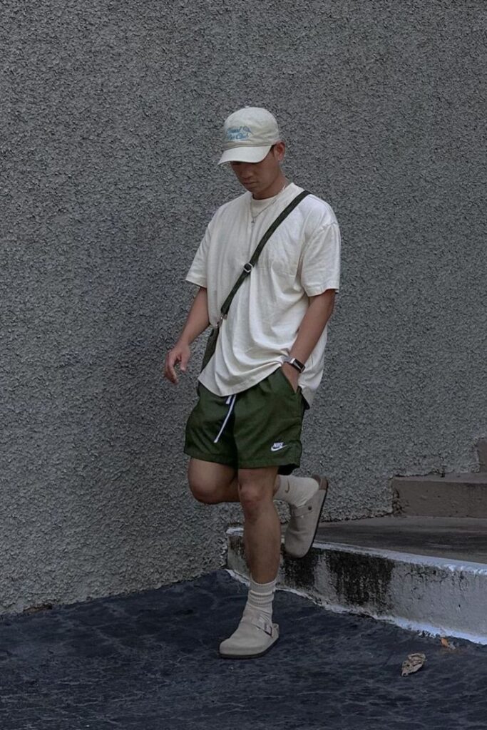 Casual street style: person in beige shirt, cap, green Nike shorts, and crossbody bag against a gray wall background.
