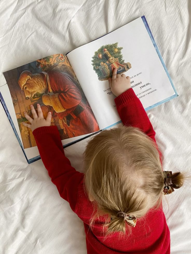 Child reading a picture book in bed, pointing at a festive illustration with two fingers.