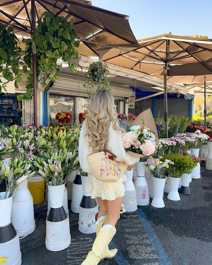 Woman in floral market holding bouquet, wearing yellow boots and a straw bag, surrounded by vibrant flowers and greenery.